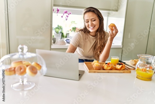 Young beautiful hispanic woman having breakfast using laptop at the kitchen