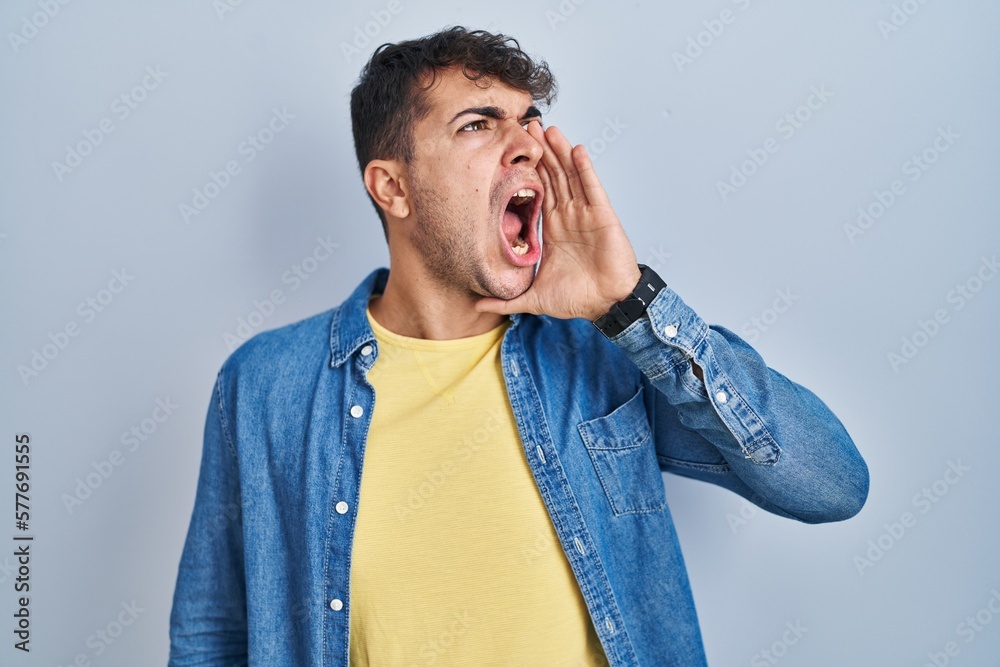 Young hispanic man standing over blue background shouting and screaming loud to side with hand on mouth. communication concept. Young hispanic man standing over blue background shouting and screaming loud to side with hand on mouth. communication concept.