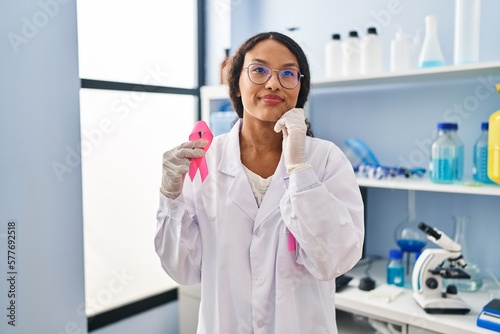 Fototapeta Naklejka Na Ścianę i Meble -  Young hispanic doctor woman working at scientist laboratory holding pink ribbon serious face thinking about question with hand on chin, thoughtful about confusing idea