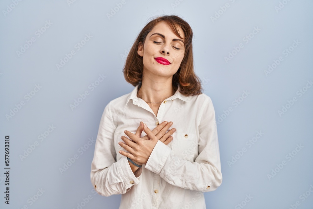 Young beautiful woman standing casual over blue background smiling with hands on chest with closed eyes and grateful gesture on face. health concept. Young beautiful woman standing casual over blue background smiling with hands on chest with closed eyes and grateful gesture on face. health concept.
