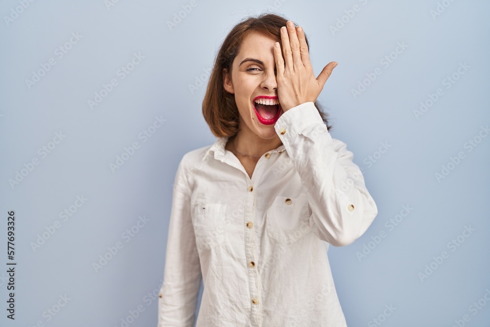 Young beautiful woman standing casual over blue background covering one eye with hand, confident smile on face and surprise emotion.