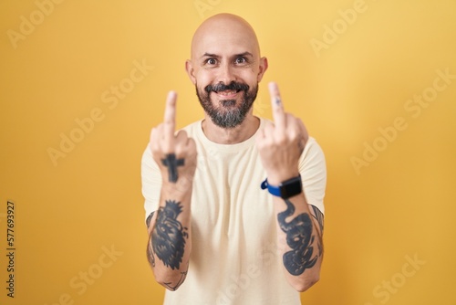 Hispanic man with tattoos standing over yellow background showing middle finger doing fuck you bad expression, provocation and rude attitude. screaming excited