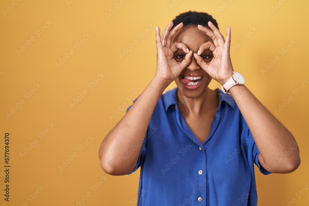 African american woman standing over yellow background doing ok gesture like binoculars sticking tongue out, eyes looking through fingers. crazy expression. African american woman standing over yellow background doing ok gesture like binoculars sticking tongue out, eyes looking through fingers. crazy expression.