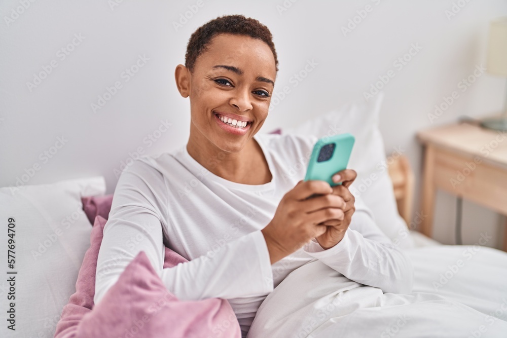African american woman using smartphone sitting on bed at bedroom African american woman using smartphone sitting on bed at bedroom