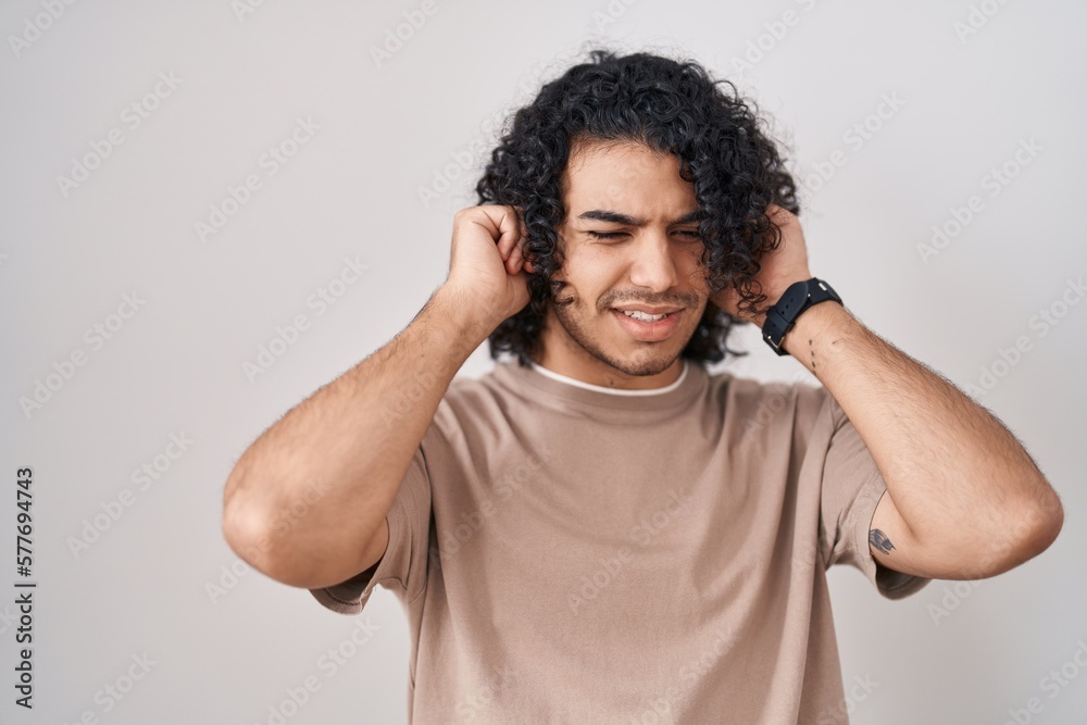 Hispanic man with curly hair standing over white background covering ears with fingers with annoyed expression for the noise of loud music. deaf concept. Hispanic man with curly hair standing over white background covering ears with fingers with annoyed expression for the noise of loud music. deaf concept.