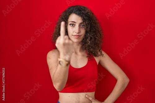 Hispanic woman with curly hair standing over red background showing middle finger, impolite and rude fuck off expression