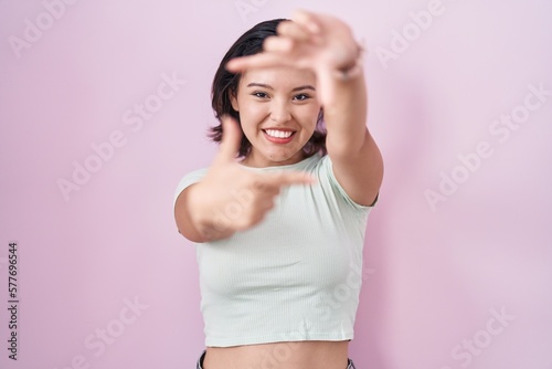 Hispanic young woman standing over pink background smiling making frame with hands and fingers with happy face. creativity and photography concept. © Krakenimages.com