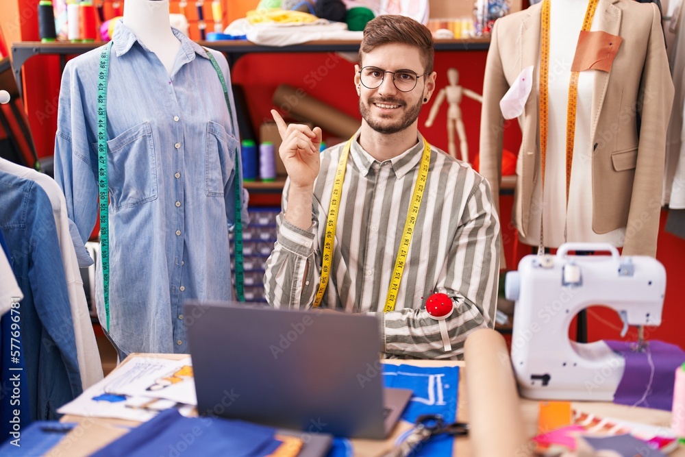 Hispanic man with beard using laptop at tailor room smiling happy pointing with hand and finger to the side