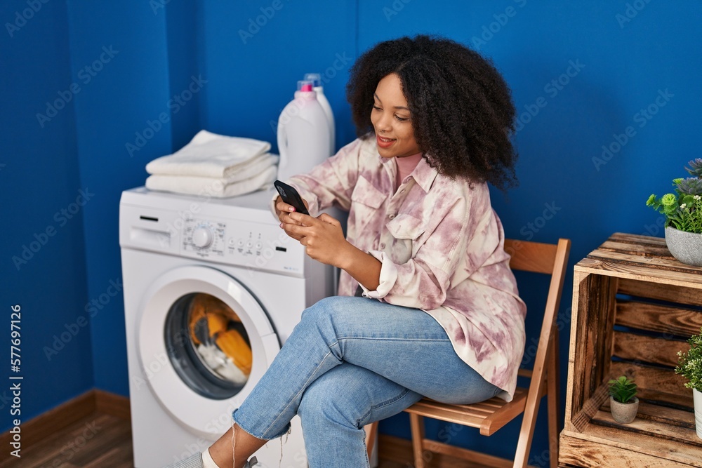 Young african american woman using smartphone waiting for washing machine at laundry Young african american woman using smartphone waiting for washing machine at laundry