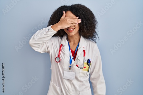 Young african american woman wearing doctor uniform and stethoscope smiling and laughing with hand on face covering eyes for surprise. blind concept.