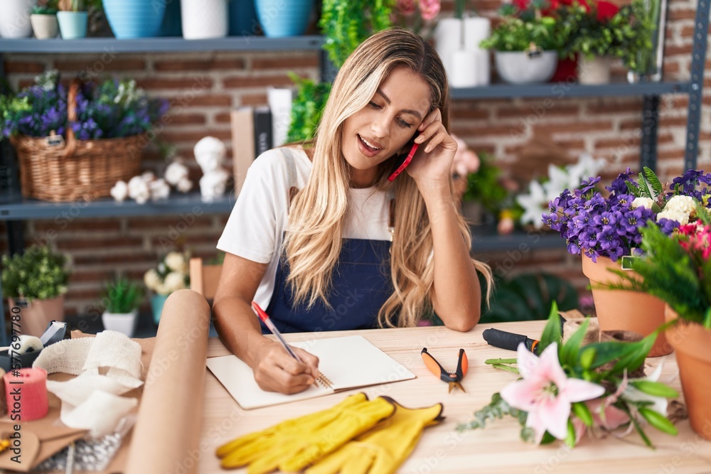 Young blonde woman florist talking on smartphone writing on notebook at florist Young blonde woman florist talking on smartphone writing on notebook at florist