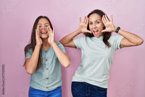 Young mother and daughter standing over pink background smiling cheerful playing peek a boo with hands showing face. surprised and exited