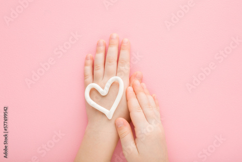 White heart shape on baby girl hand on light pink table background. Pastel color. Closeup. Point of view shot. Care about child body skin. Top down view.