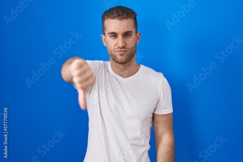 Young caucasian man standing over blue background looking unhappy and angry showing rejection and negative with thumbs down gesture. bad expression.