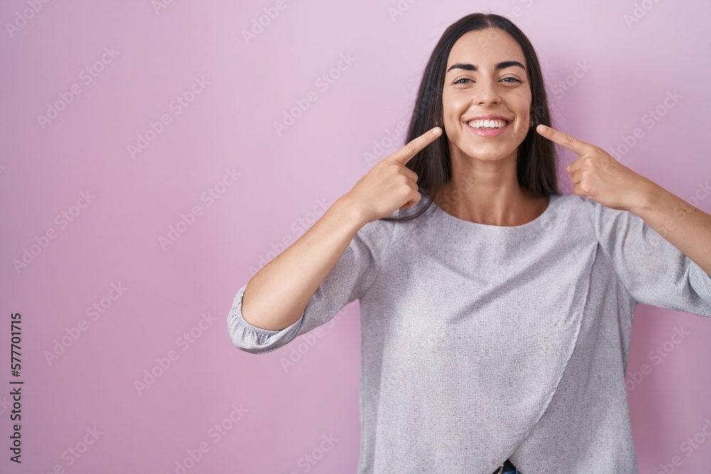 Young brunette woman standing over pink background smiling cheerful showing and pointing with fingers teeth and mouth. dental health concept. Young brunette woman standing over pink background smiling cheerful showing and pointing with fingers teeth and mouth. dental health concept.