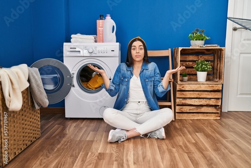 Hispanic woman doing laundry sitting on the floor clueless and confused expression with arms and hands raised. doubt concept.