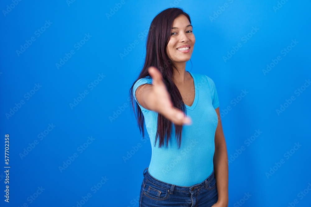 Young hispanic woman standing over blue background smiling friendly offering handshake as greeting and welcoming. successful business. Young hispanic woman standing over blue background smiling friendly offering handshake as greeting and welcoming. successful business.