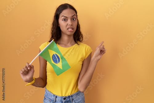 Young hispanic woman holding brazil flag pointing aside worried and nervous with forefinger  concerned and surprised expression