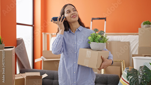 Young beautiful hispanic woman listening to voice message with smartphone holding a box at new home