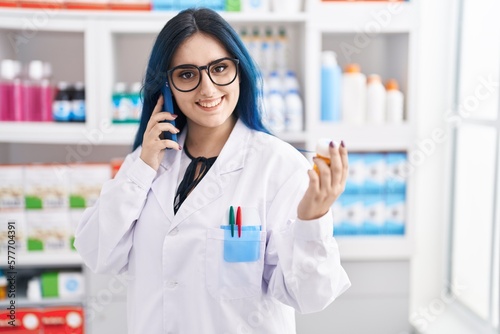 Young caucasian woman pharmacist holding pills bottle talking on smartphone at pharmacy