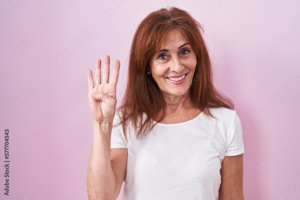 Middle age woman standing over pink background showing and pointing up with fingers number four while smiling confident and happy. Middle age woman standing over pink background showing and pointing up with fingers number four while smiling confident and happy.