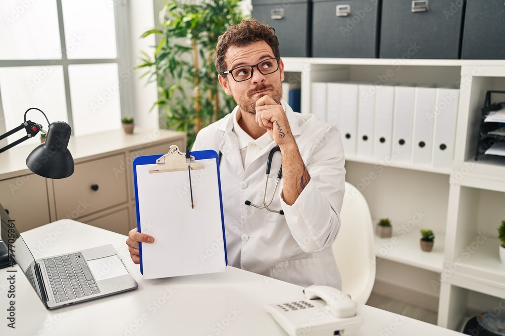 Young hispanic man wearing doctor stethoscope holding clipboard serious face thinking about question with hand on chin, thoughtful about confusing idea