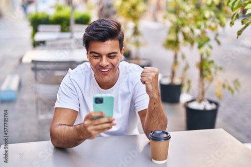 Young hispanic man using smartphone drinking coffee at coffee shop terrace