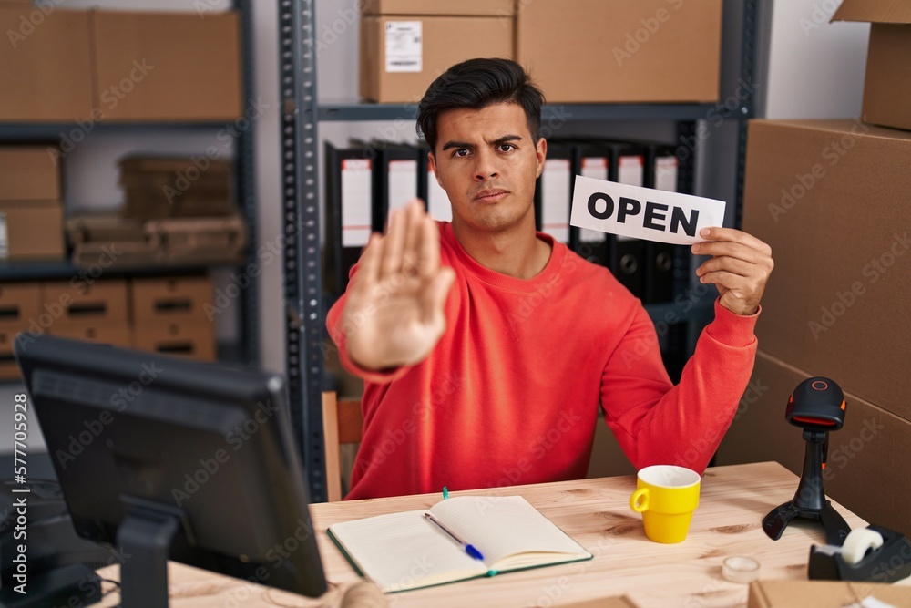 Hispanic man working at small business ecommerce holding open banner with open hand doing stop sign with serious and confident expression, defense gesture