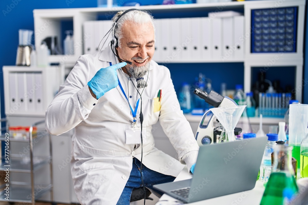 Middle age man with grey hair working at scientist laboratory doing video call smiling happy pointing with hand and finger