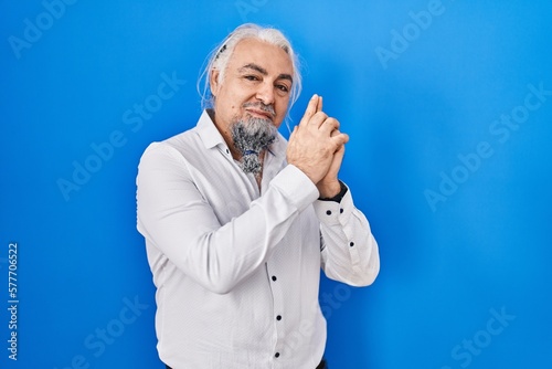 Middle age man with grey hair standing over blue background holding symbolic gun with hand gesture, playing killing shooting weapons, angry face