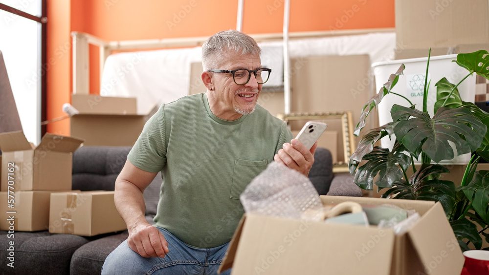 Middle age grey-haired man unpacking cardboard box using smartphone at new home Middle age grey-haired man unpacking cardboard box using smartphone at new home