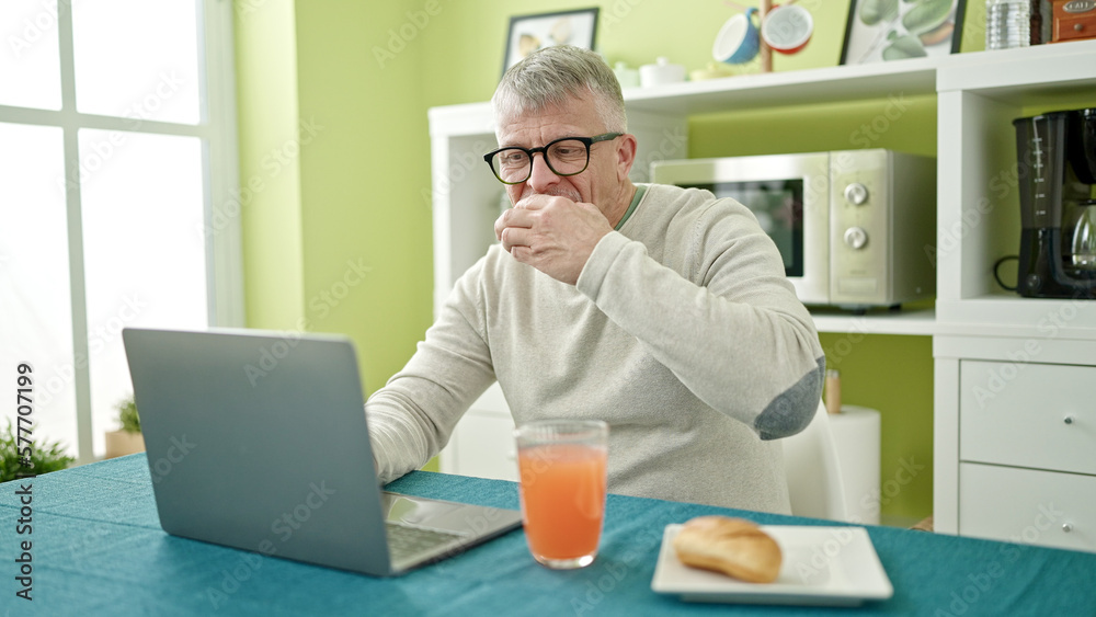 Middle age grey-haired man having breakfast using laptop at home