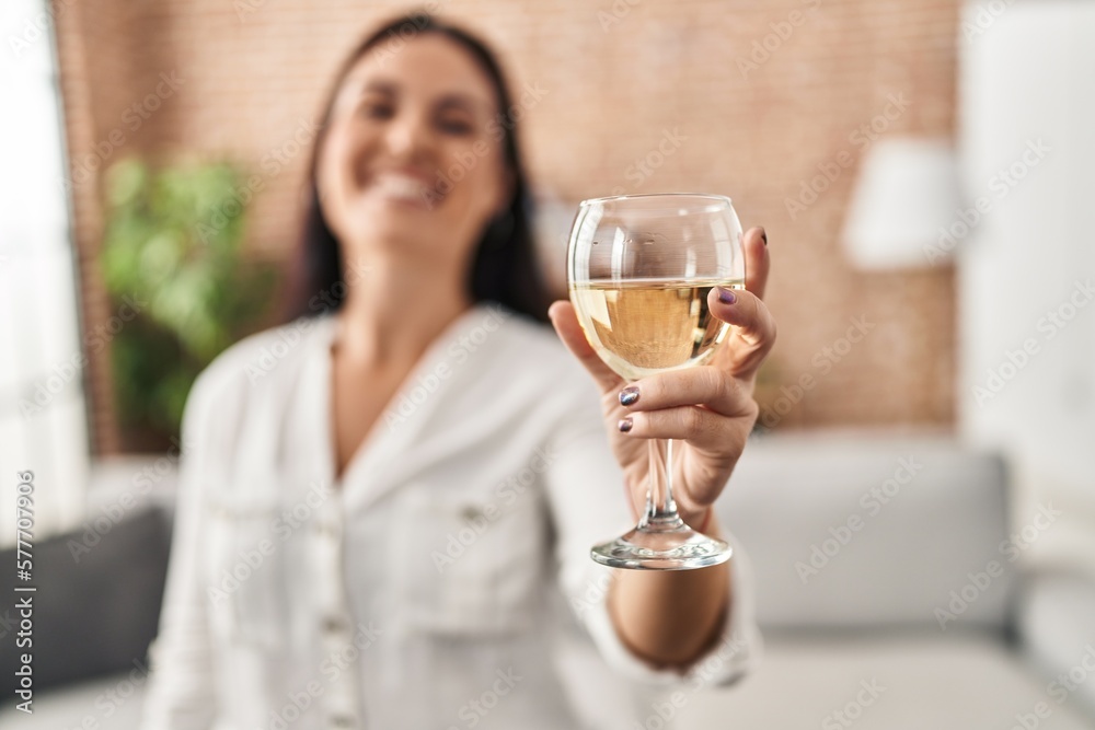 Young beautiful hispanic woman drinking glass of wine standing at home