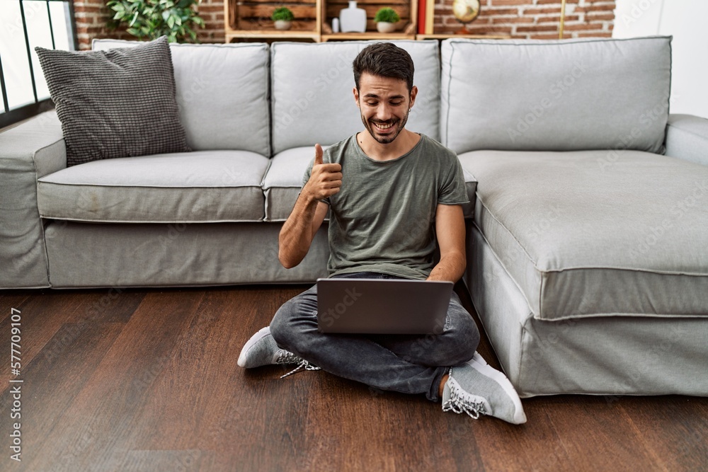 Young hispanic man using laptop sitting on the at home smiling happy and positive, thumb up doing excellent and approval sign