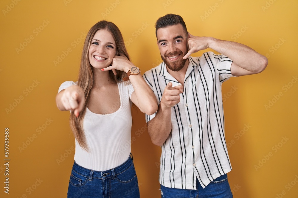 Young couple standing over yellow background smiling doing talking on the telephone gesture and pointing to you. call me. Young couple standing over yellow background smiling doing talking on the telephone gesture and pointing to you. call me.