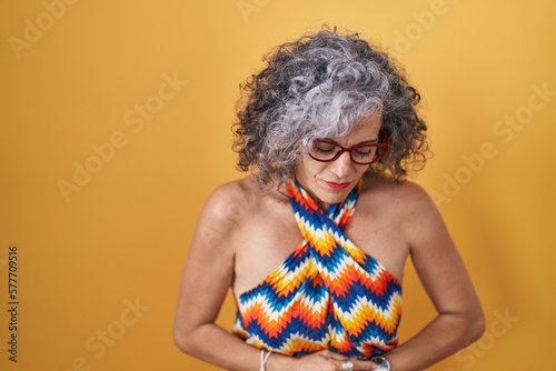 Middle age woman with grey hair standing over yellow background with hand on stomach because indigestion, painful illness feeling unwell. ache concept.