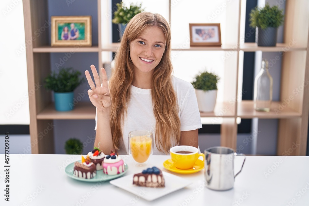 Young caucasian woman eating pastries t for breakfast showing and pointing up with fingers number three while smiling confident and happy.