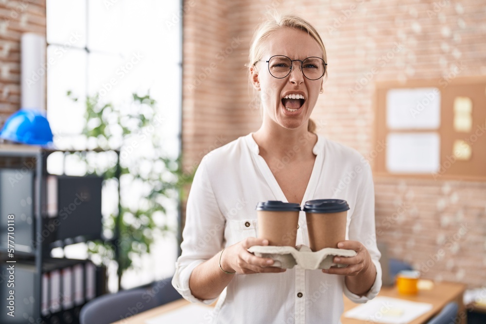Young caucasian woman working at the office holding coffee cups angry and mad screaming frustrated and furious, shouting with anger. rage and aggressive concept.
