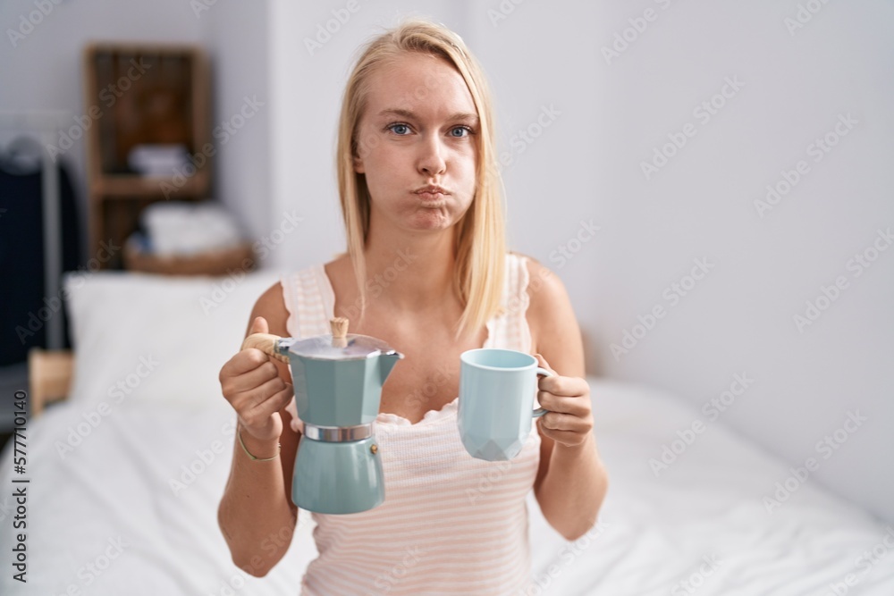 Young caucasian woman drinking coffee on the bed puffing cheeks with funny face. mouth inflated with air, catching air. Young caucasian woman drinking coffee on the bed puffing cheeks with funny face. mouth inflated with air, catching air.