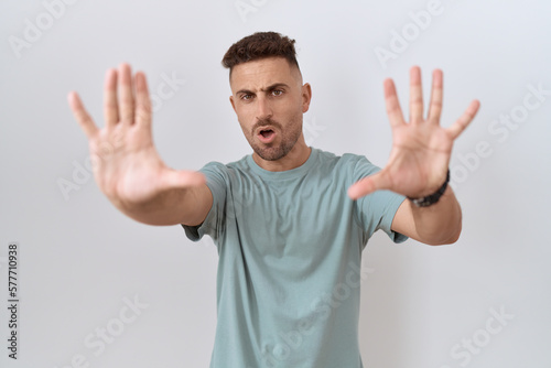 Hispanic man with beard standing over white background doing stop gesture with hands palms, angry and frustration expression