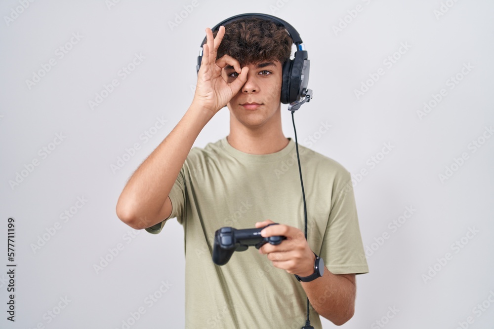 Hispanic teenager playing video game holding controller doing ok gesture with hand smiling, eye looking through fingers with happy face.