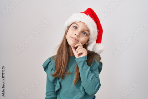 Little caucasian girl wearing christmas hat smiling looking confident at the camera with crossed arms and hand on chin. thinking positive.