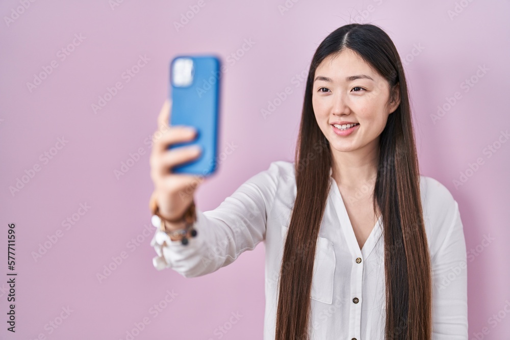 Chinese young woman taking a selfie photo with smartphone looking positive and happy standing and smiling with a confident smile showing teeth Chinese young woman taking a selfie photo with smartphone looking positive and happy standing and smiling with a confident smile showing teeth