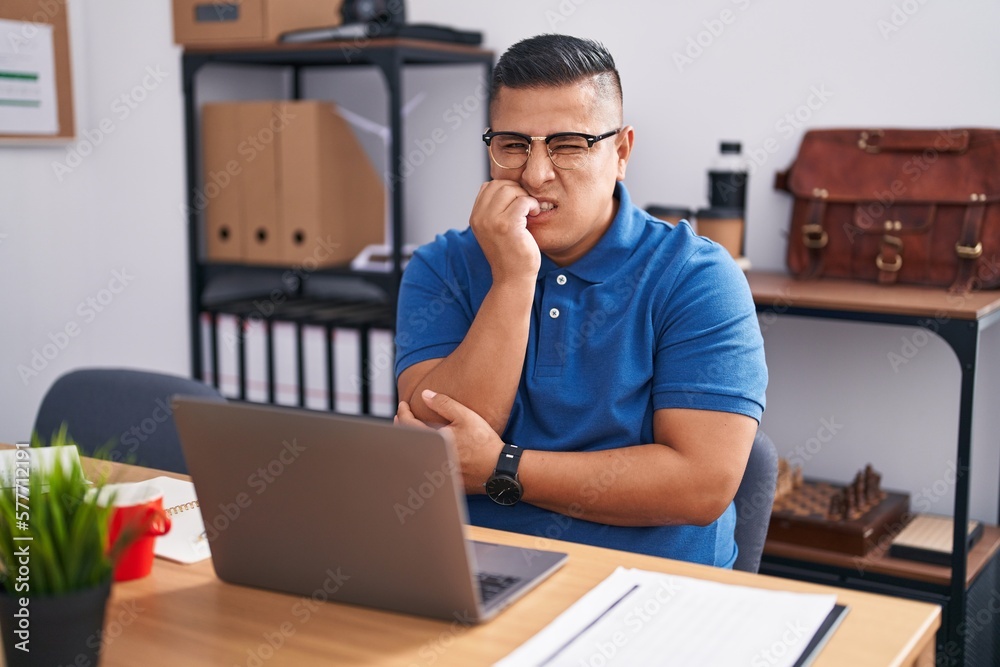 Young hispanic man working at the office with laptop looking stressed and nervous with hands on mouth biting nails. anxiety problem. Young hispanic man working at the office with laptop looking stressed and nervous with hands on mouth biting nails. anxiety problem.