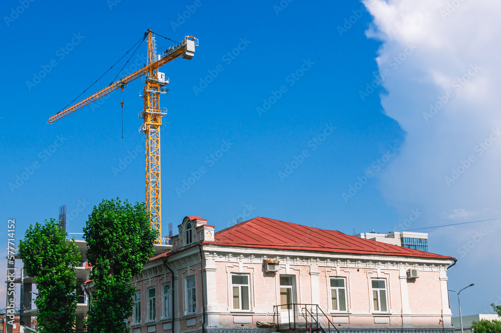 Behind an old house built in the 19th century there is a crane that lifts the load during the construction of a new high-rise building.