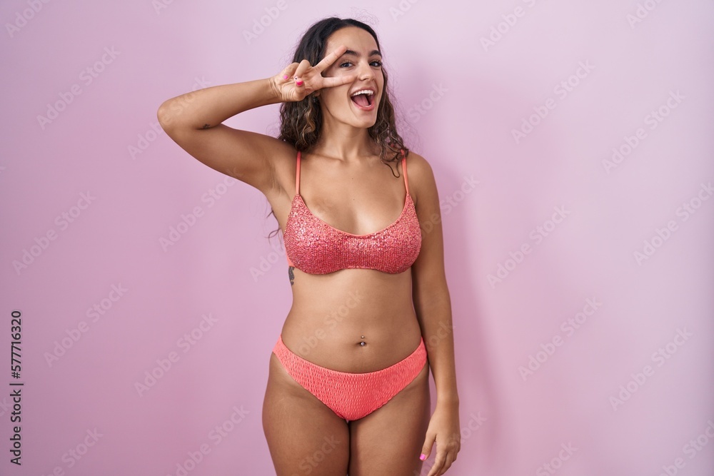 Fototapeta premium Young hispanic woman wearing lingerie over pink background doing peace symbol with fingers over face, smiling cheerful showing victory