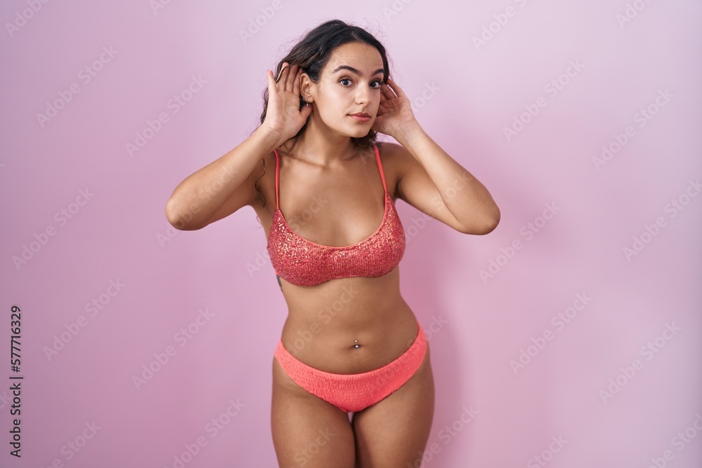 Young hispanic woman wearing lingerie over pink background trying to hear both hands on ear gesture, curious for gossip. hearing problem, deaf