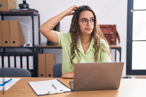 Young hispanic woman working at the office wearing glasses confuse and wonder about question. uncertain with doubt, thinking with hand on head. pensive concept.