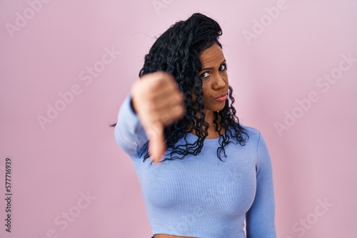 Middle age hispanic woman standing over pink background looking unhappy and angry showing rejection and negative with thumbs down gesture. bad expression.