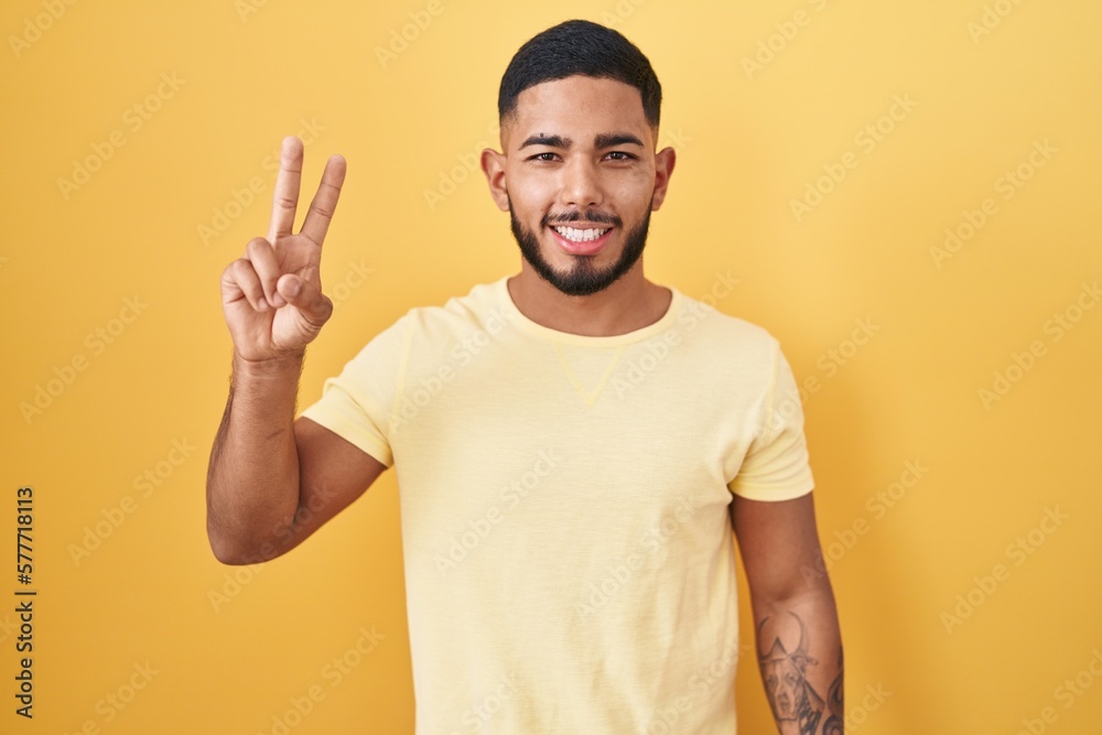 Young hispanic man standing over yellow background smiling looking to the camera showing fingers doing victory sign. number two. Young hispanic man standing over yellow background smiling looking to the camera showing fingers doing victory sign. number two.